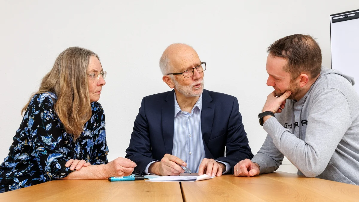 Drei Personen sitzen am Tisch und führen ein intensives Beratungsgespräch, bei dem gemeinsam mögliche Vorteile und Entscheidungen besprochen werden.